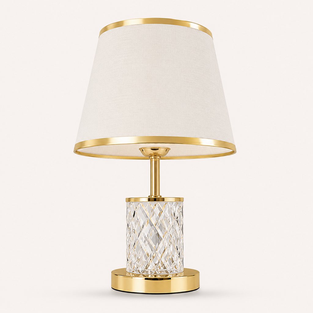 A modern table lamp with a tapered off-white fabric shade, polished gold metal trim, a clear faceted crystal cylinder base, and a round gold metal base, shown against a light beige background.