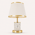 A modern table lamp with a tapered off-white fabric shade, polished gold metal trim, a clear faceted crystal cylinder base, and a round gold metal base, shown against a light beige background.