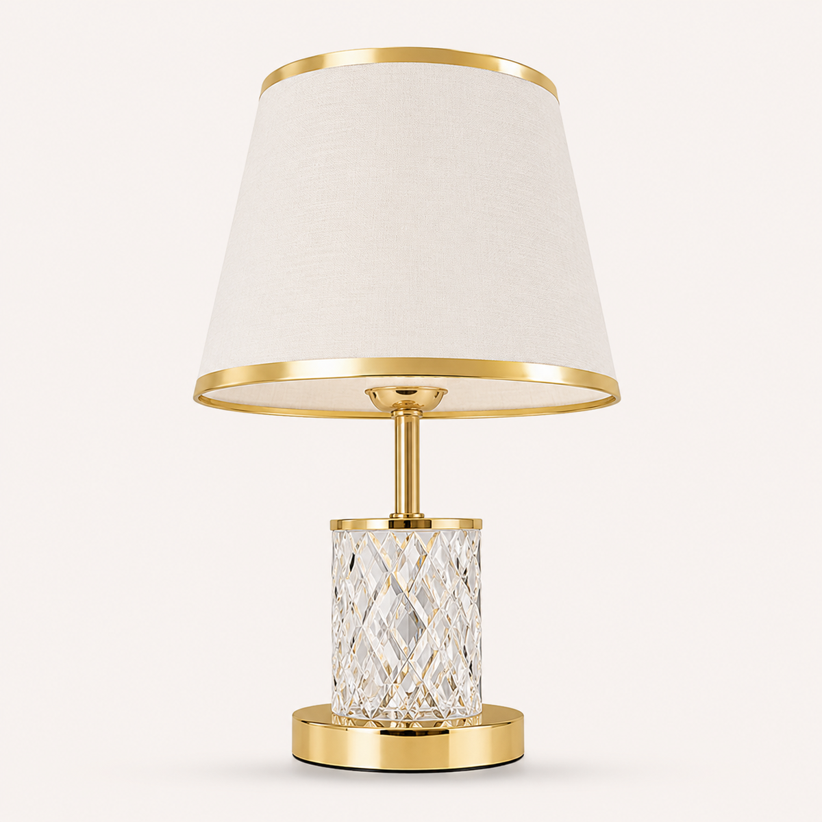 A modern table lamp with a tapered off-white fabric shade, polished gold metal trim, a clear faceted crystal cylinder base, and a round gold metal base, shown against a light beige background.