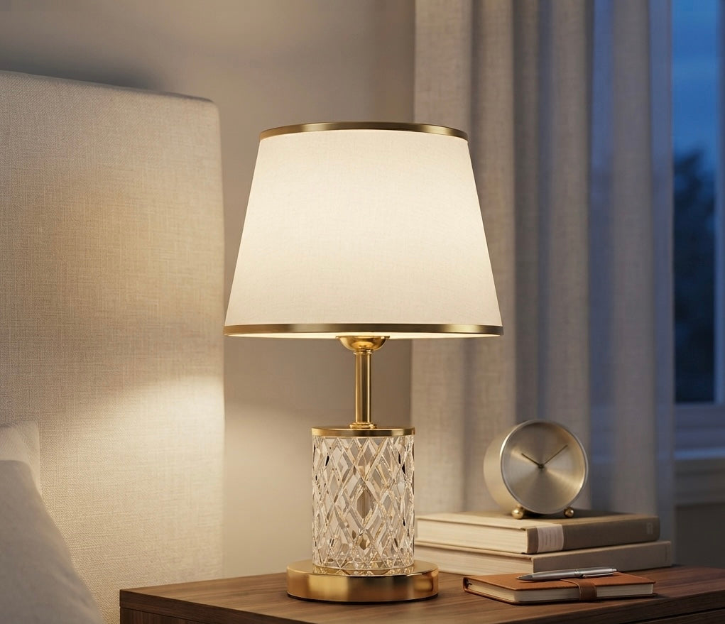 A table lamp with a warm white fabric shade, gold-toned metal trim and stem, and a faceted clear crystal base, shown lit on a wooden bedside table next to books and a small clock.