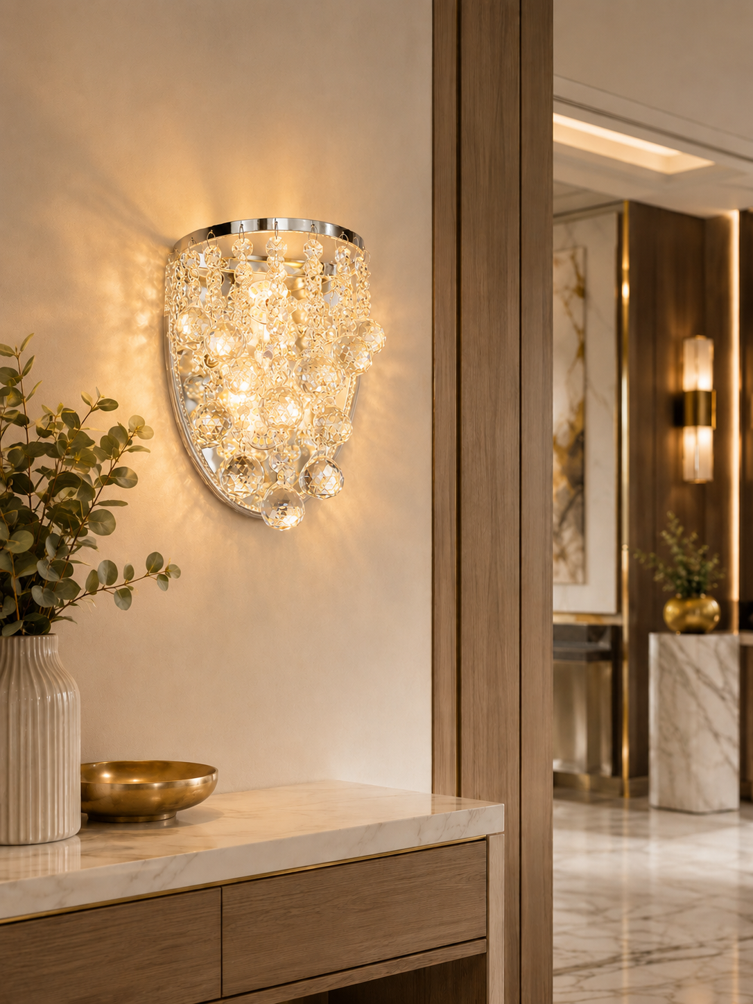 A modern wall sconce with a curved silver metal frame and cascading clear crystal droplets, illuminated with warm light on a beige wall above a marble console table.