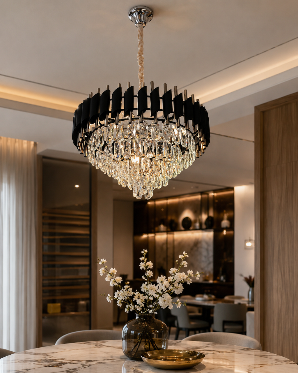 A modern circular chandelier with a black fluted outer ring and hanging clear crystal prisms, suspended over a marble dining table in a contemporary interior.